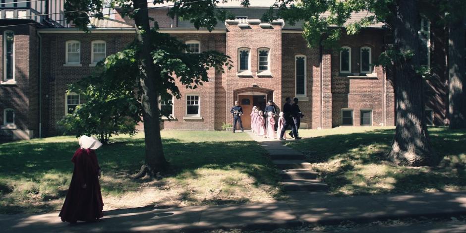 Ofglen and Offred walk down the street as some guards lead a group of schoolchildren outside.