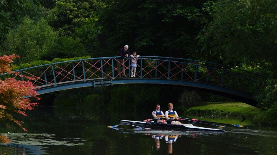 Two rowers continue underneath a bridge where Whispers is standing with his daughter.