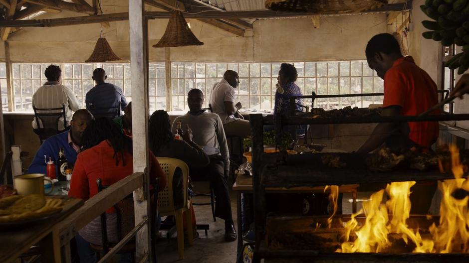 Capheus and Zakia sit at the table along the window while someone prepares food in the foreground.