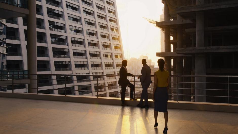 Kala walks out onto the balcony to where Ajay and Rajan are looking out at the construction lit by the sunset.