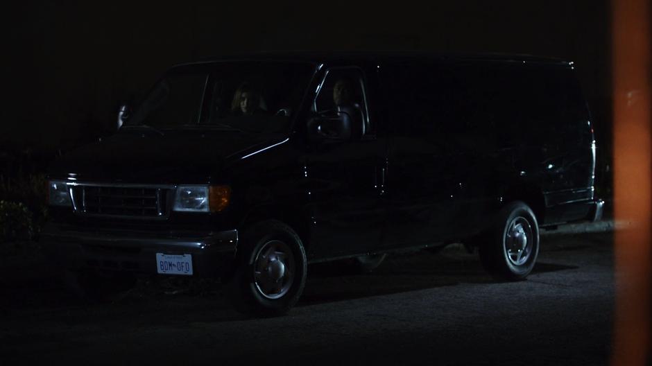 Dinah and Curtis watch Derek Sampson pick up a bunch of big guns from their van across from a sketchy warehouse.