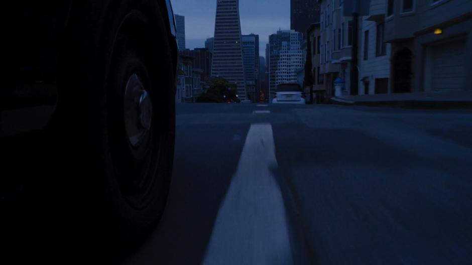 Bendix's car drives through the intersection as Amanita drives down the hill ahead.