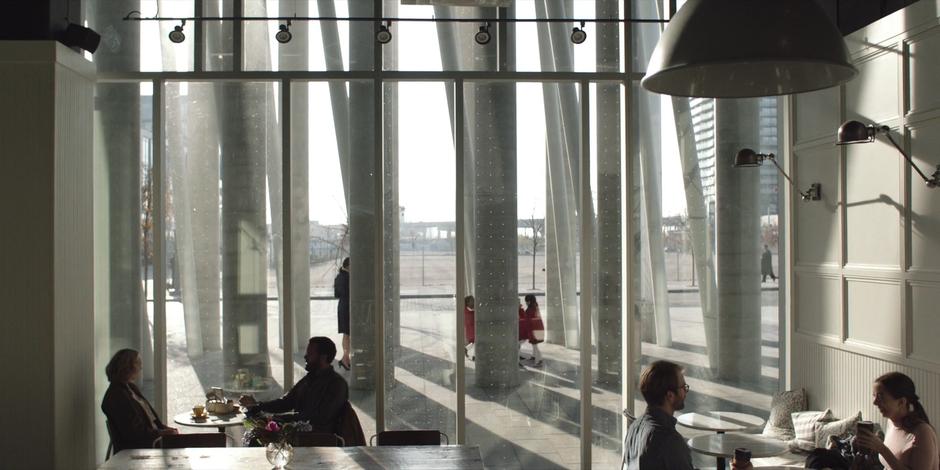 June and Luke sit at a table drinking their coffee while children in red outfits run around outside.