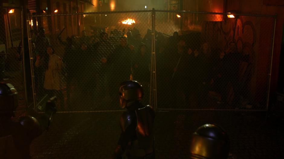 A group of civilians push against the fence while several Daxamite soldier point guns at them.