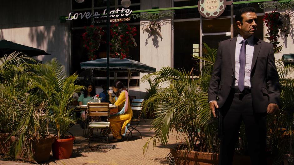 Kala and Priya sit at a table in the cafe drinking coffee while their guard stands on the street looking around.