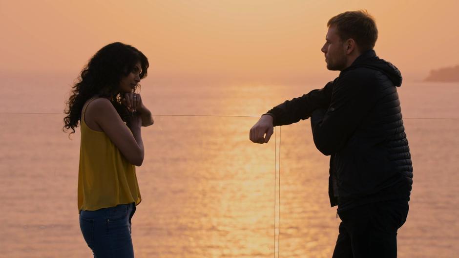 Kala leans on the glass railing and faces Wolfgang in front of the sunset.