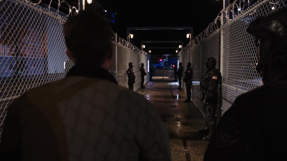 Adrian Chase is escorted by several A.R.G.U.S. agents down a fenced-in corridor towards a landing helipad.