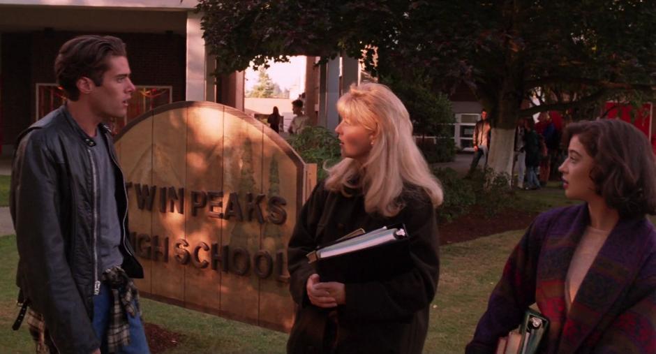 Laura talks with Bobby in front of the school sign while Donna watches.