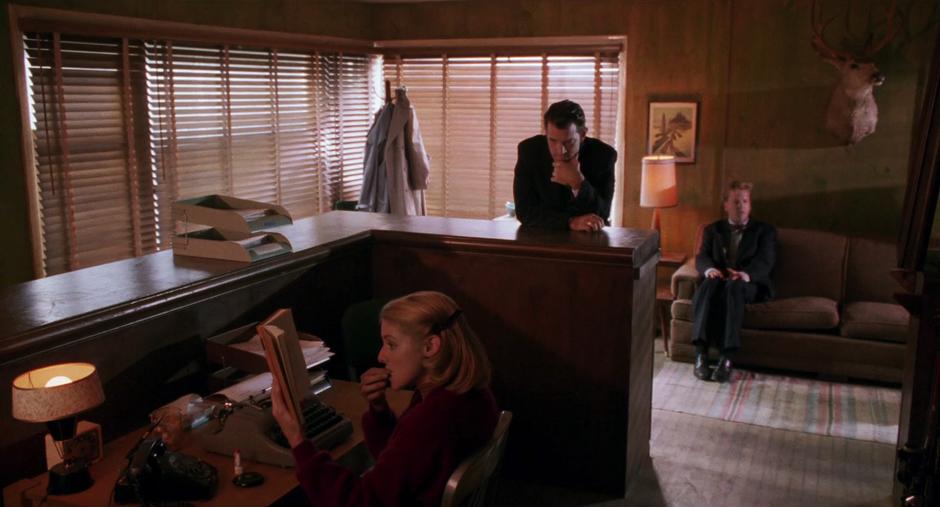 The Sheriff's Department receptionist reads a book at her desk while Chet leans impatiently against the counter and Sam sits awkwardly on the couch.