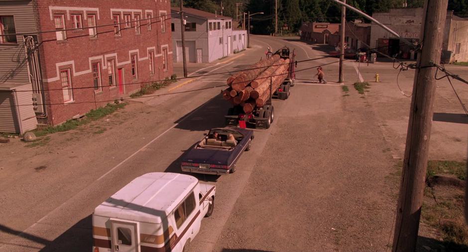 A log truck, Leland & Laura's car, and the One-Armed Man's truck wait at the intersection as a woman helps an old man with a walker cross the street.