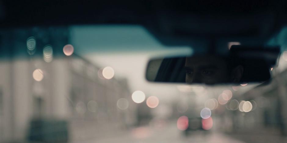 Nick's eyes are visible in the rearview mirror as he drives down the street.