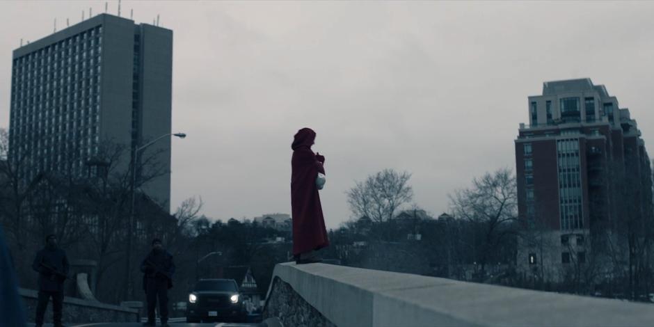 Janine stands on the edge of the bridge holding her child while several guards look on.