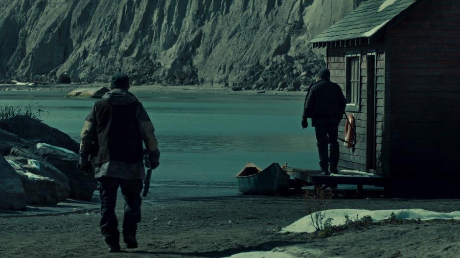 A guard walks up to the boathouse where another waits looking off across the water.