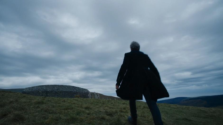 The Doctor walks up the hill towards the stone cairn.