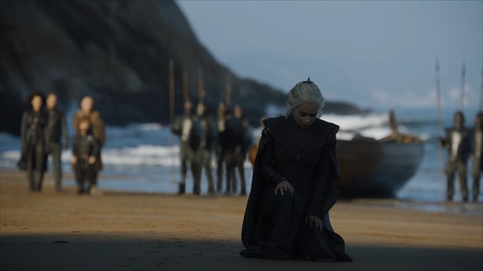 Dany kneels down the beach to touch the soil of Westeros while her guards and advisors stand in the background.