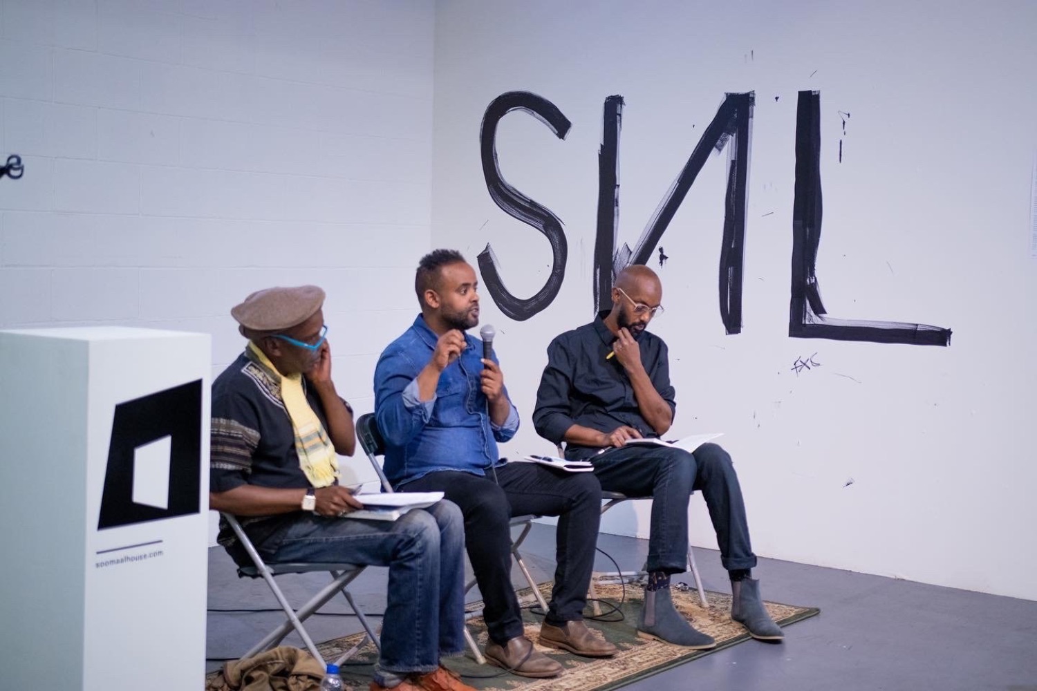 Three men sit in folding chairs and speak into a microphone next to "SNL" painted on white wall