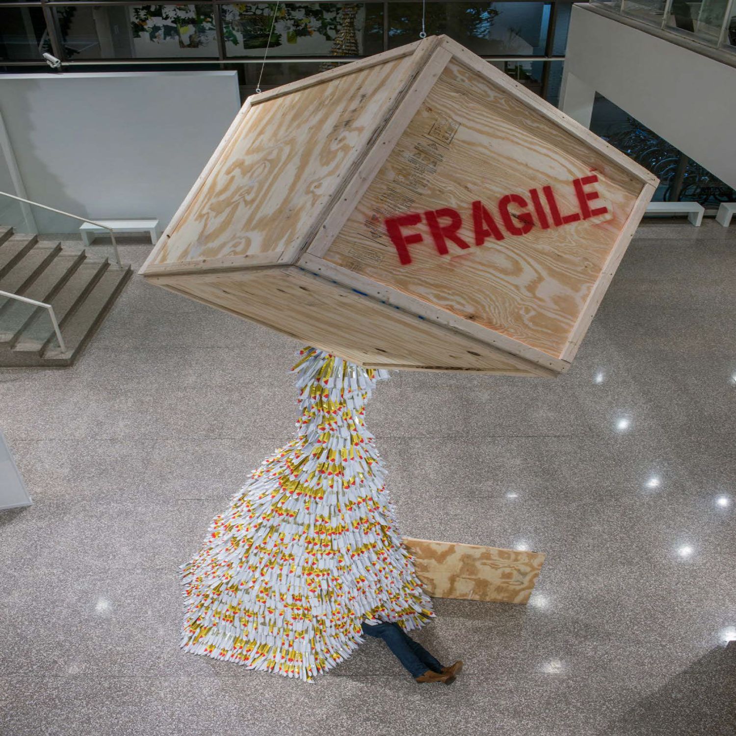 Wooden crate marked "Fragile" hangs in a gallery, a waterfall of gold and white paper spilling out onto the floor with mannequin legs sticking out from beneath the pile
