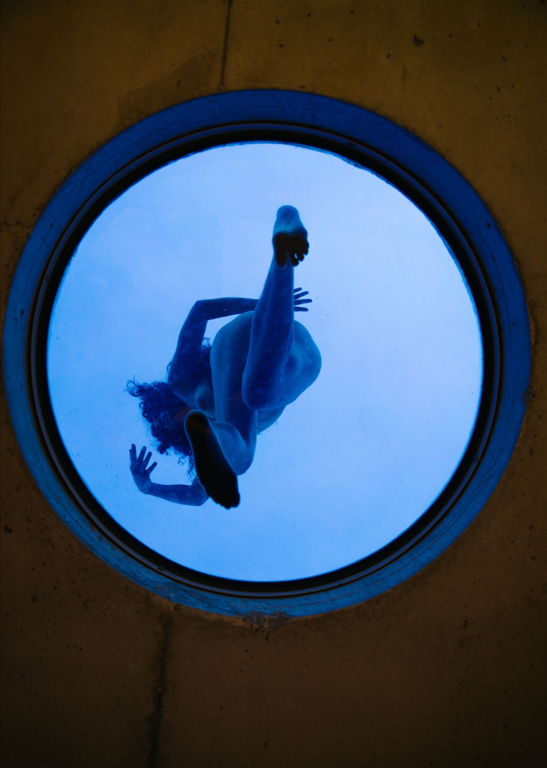 Nude woman lit in blue and seen from below standing on a circular window