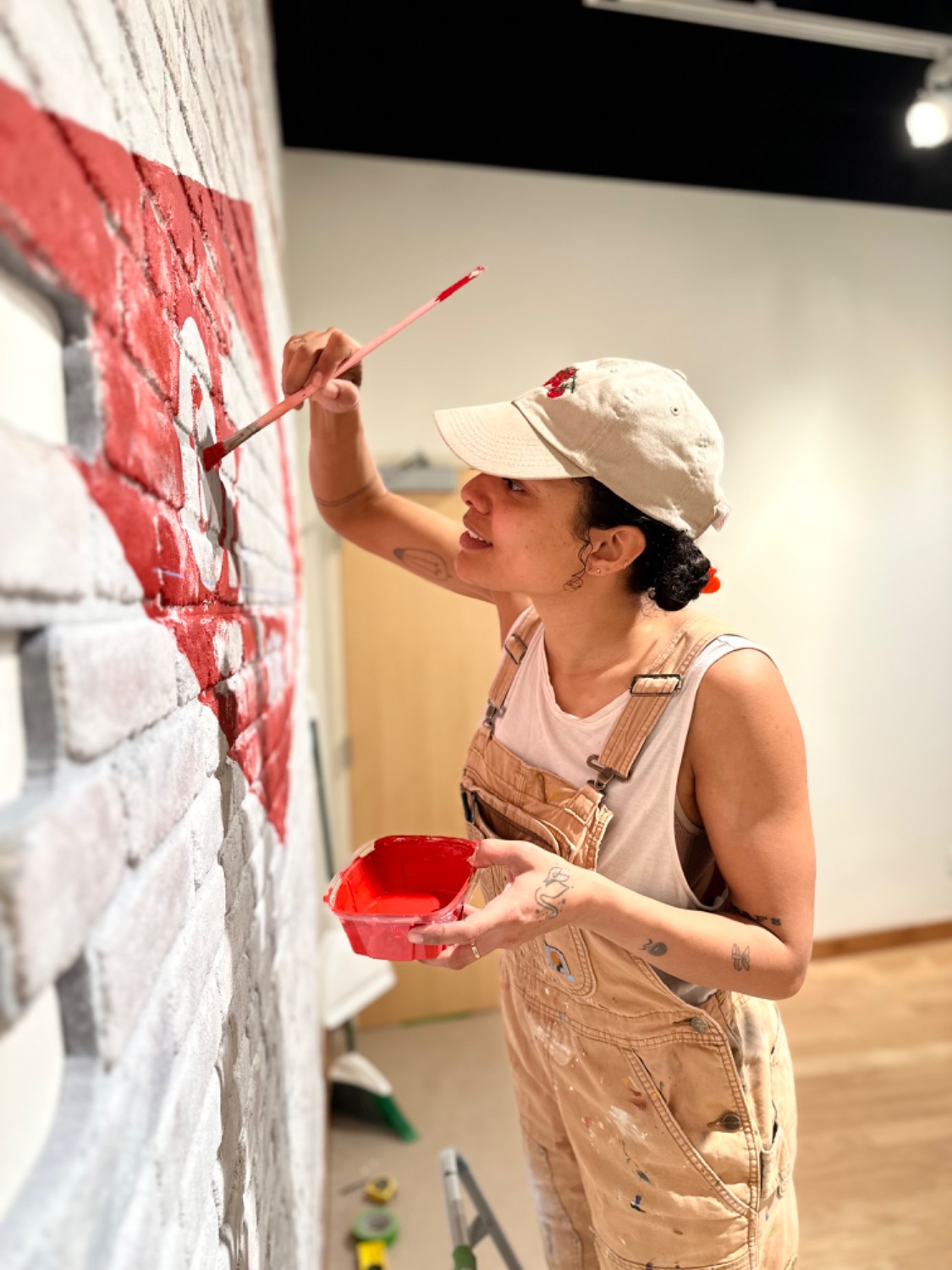 Smiling woman in hat and overalls paints a red and white sign on a brick wall.
