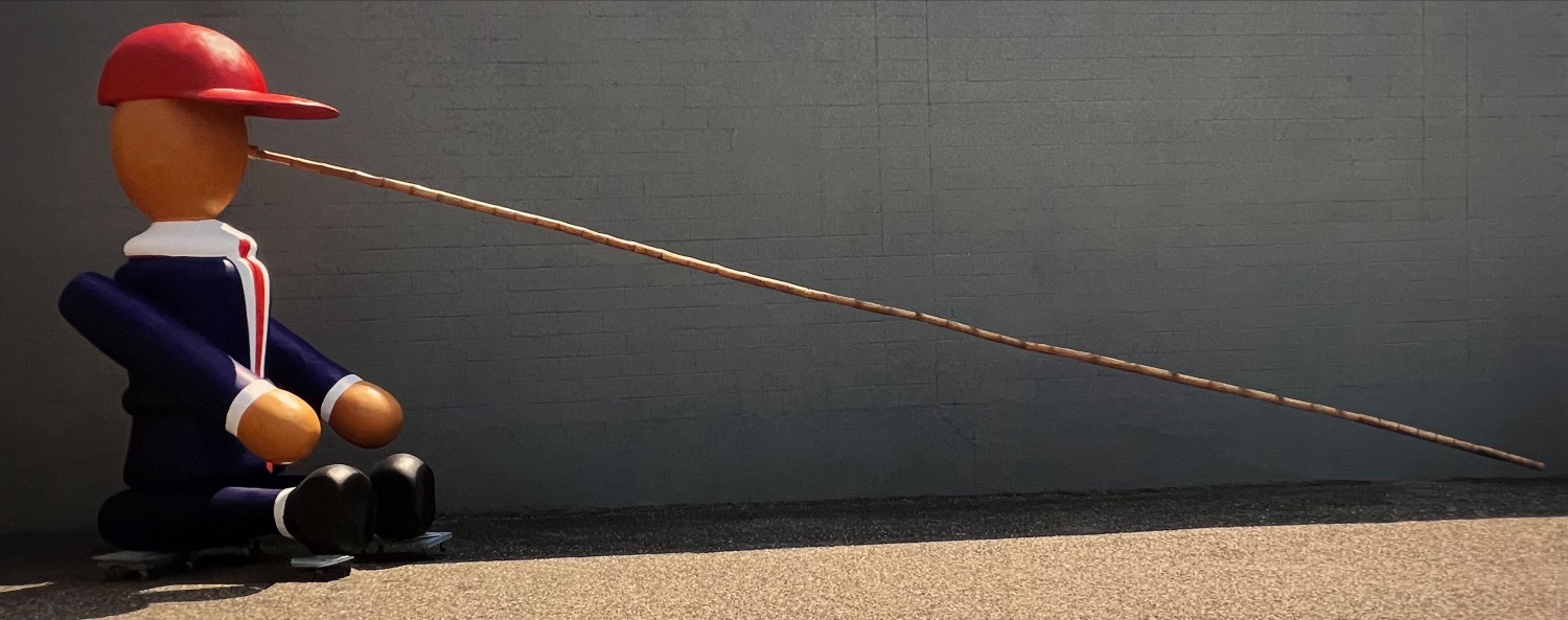 Outdoor sculpture of Pinocchio wearing red baseball hat with long bamboo nose