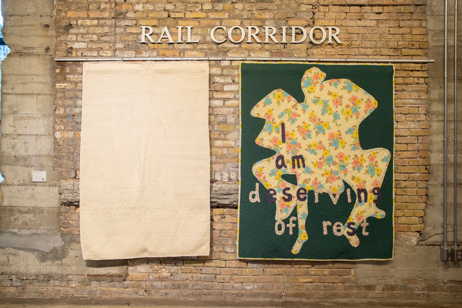 Two quilts – one white, the other green with a blob shape in flower patterned fabric and the phrase "I am deserving of rest" hang on a brick wall under metal letters "Rail corridor".