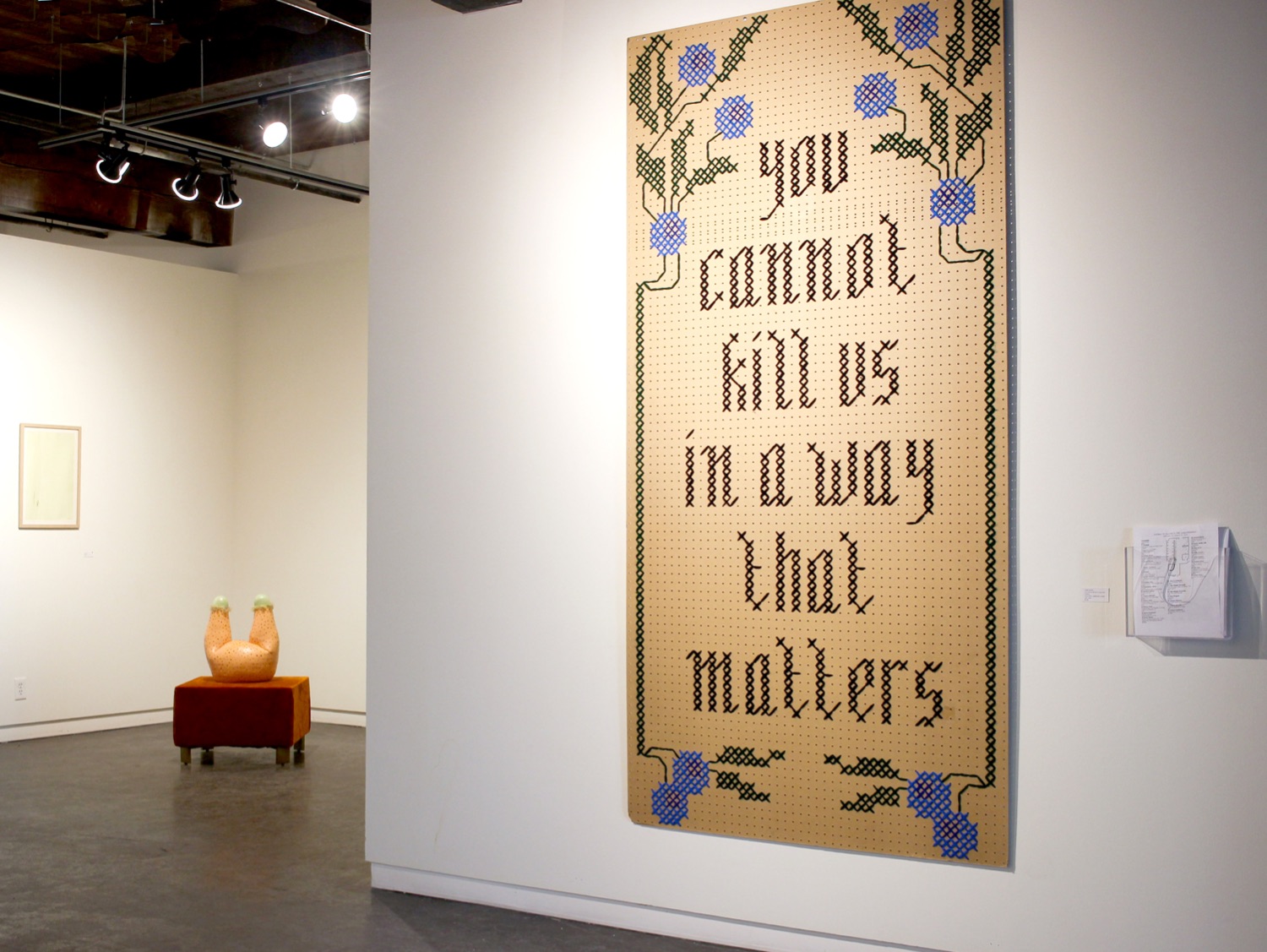 Large cross stitch artwork in a gallery reading "you cannot kill us in a way that matters"