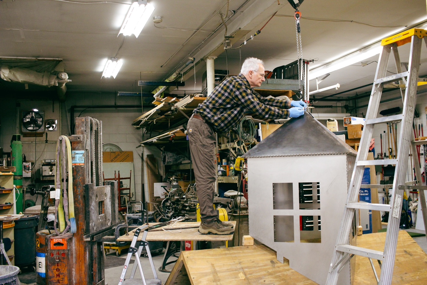 Man stands on a table adjusting a sculpture of a metal house.