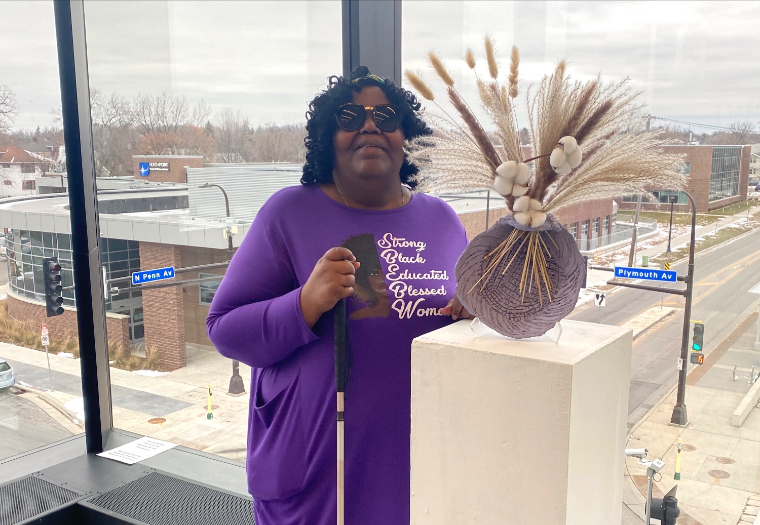 A Black woman wearing a purple shirt and sunglasses stands next to a ceramic vase in front of a second story window