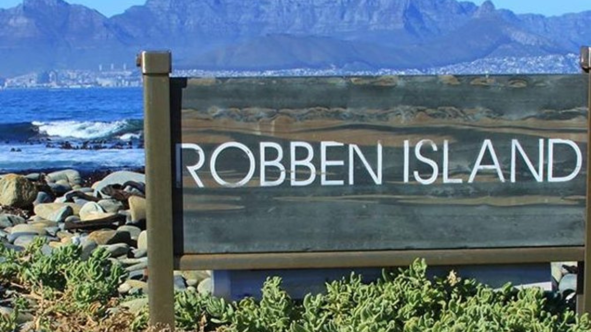 Wooden sign reading "Robben Island" with ocean waves and rocky shoreline in the foreground, and the cityscape and mountains of Cape Town, South Africa visible in the background. Wooden sign reading "Robben Island" with ocean waves and rocky shoreline in the foreground, and the cityscape and mountains of Cape Town, South Africa visible in the background.