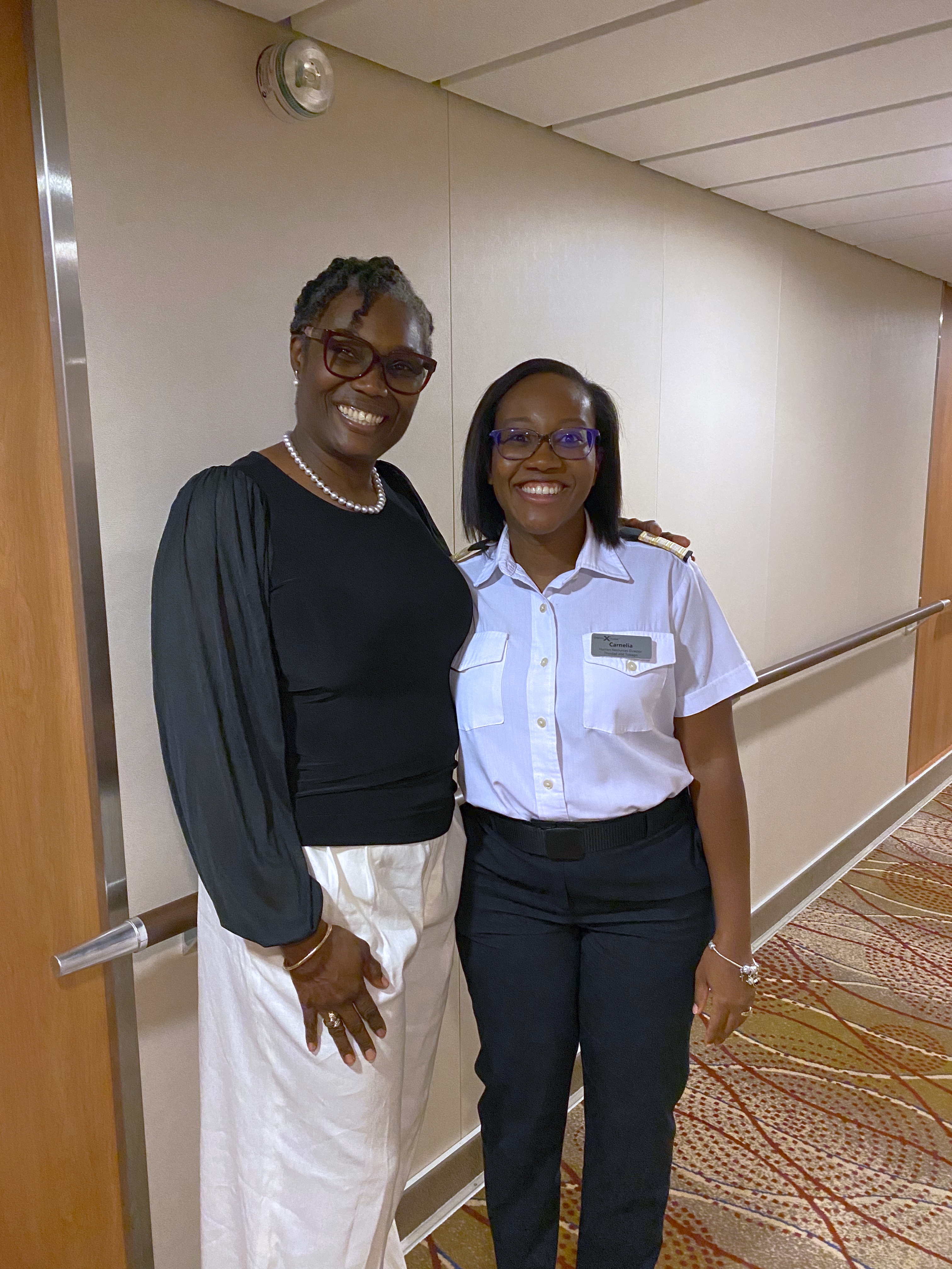 Two women smiling and posing together in a cruise ship hallway—one wearing a black top with white pants and pearls, the other in a white short-sleeve uniform with dark pants and a name tag. Two women smiling and posing together in a cruise ship hallway—one wearing a black top with white pants and pearls, the other in a white short-sleeve uniform with dark pants and a name tag.
