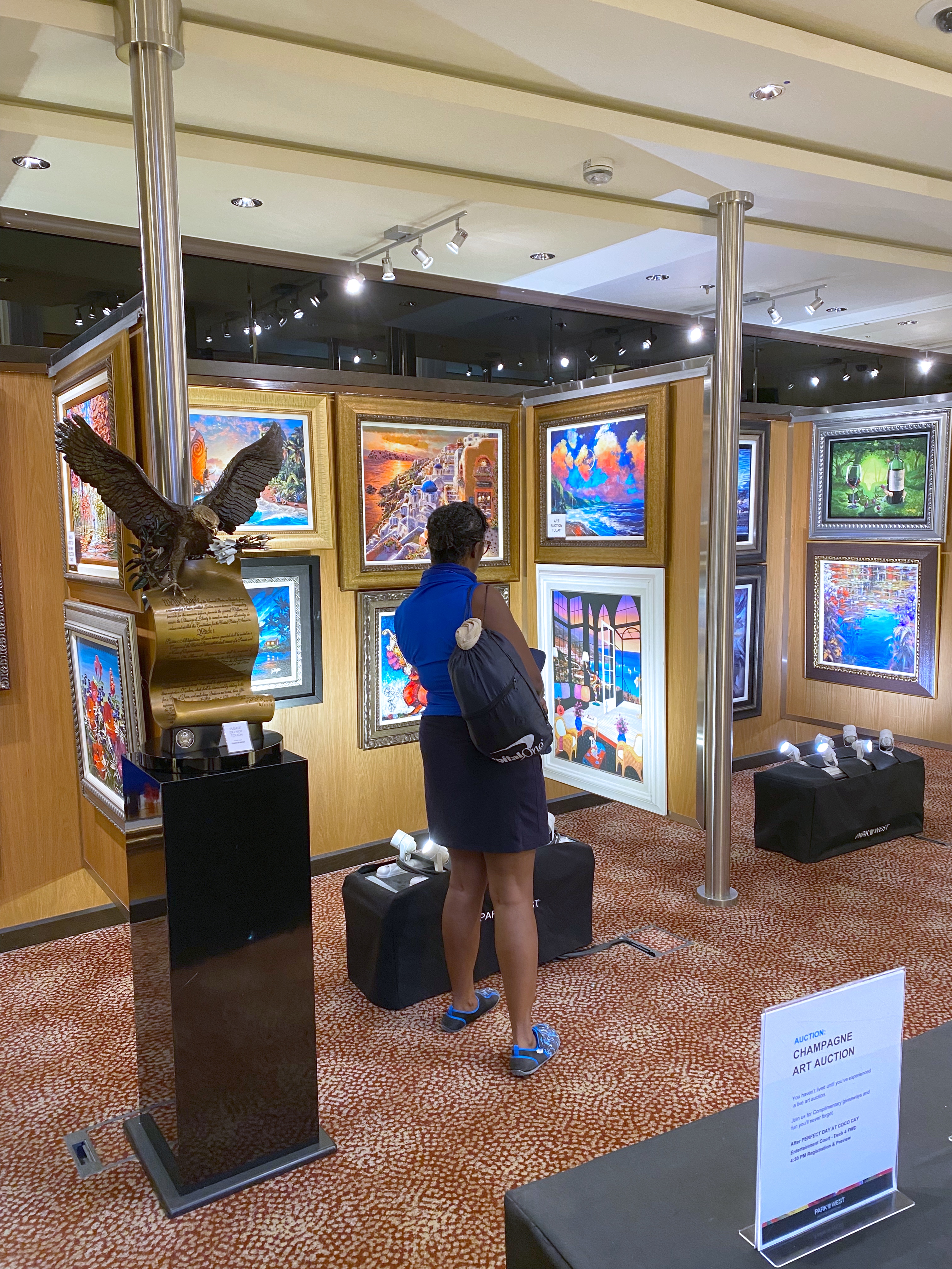 A woman in a blue top and navy skirt with a crossbody bag browses colorful framed paintings at an onboard art gallery during a cruise. The gallery is well-lit with track lighting and features vibrant works from artists like Romero Britto. A sign in the foreground announces a “Champagne Art Auction.” A woman in a blue top and navy skirt with a crossbody bag browses colorful framed paintings at an onboard art gallery during a cruise. The gallery is well-lit with track lighting and features vibrant works from artists like Romero Britto. A sign in the foreground announces a “Champagne Art Auction.”