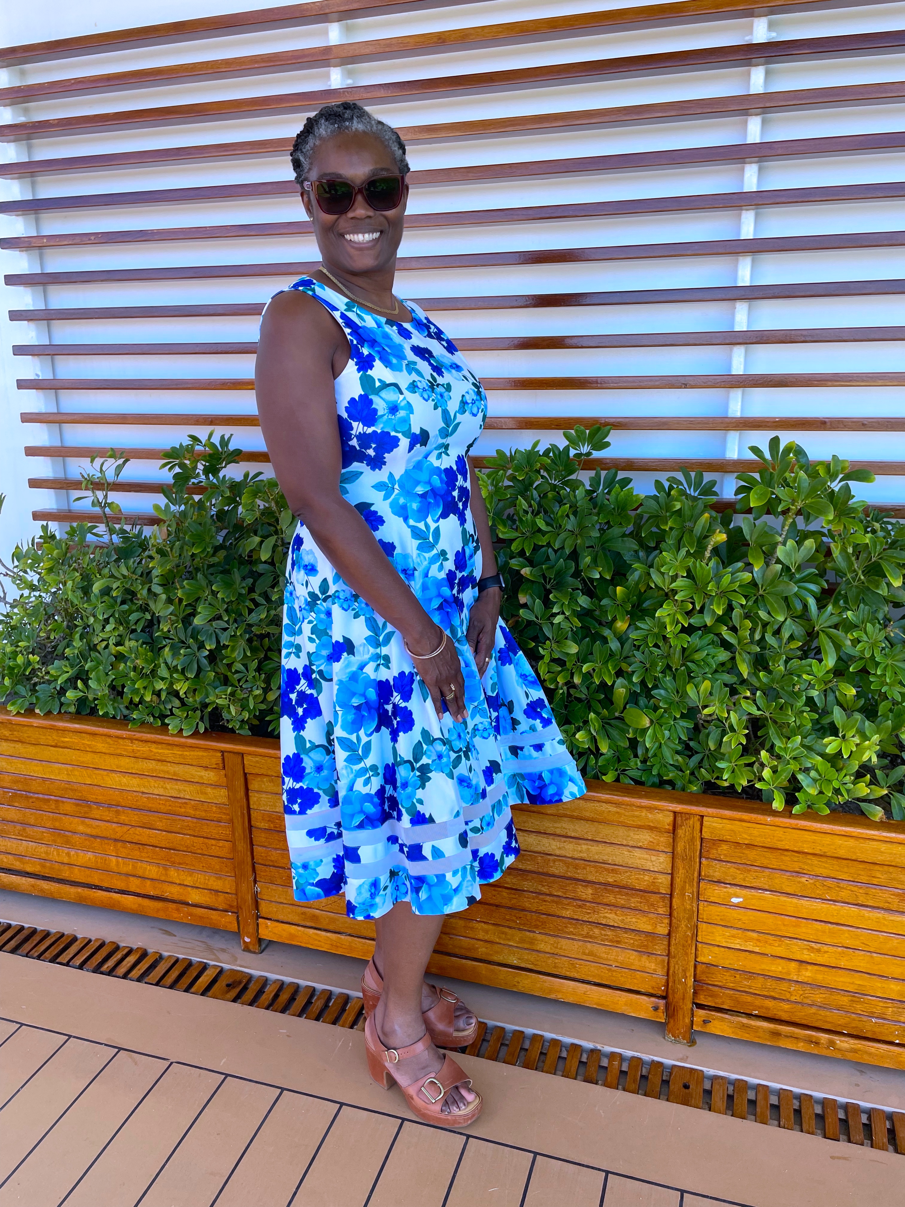 A woman in a blue and white floral dress posing on the cruise ship deck, standing in front of a planter with lush greenery and wooden slats, smiling warmly. A woman in a blue and white floral dress posing on the cruise ship deck, standing in front of a planter with lush greenery and wooden slats, smiling warmly.