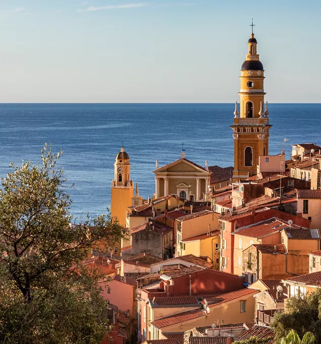 View of Menton, France, with colorful terracotta rooftops, historic church towers, and the blue Mediterranean Sea in the background. View of Menton, France, with colorful terracotta rooftops, historic church towers, and the blue Mediterranean Sea in the background.