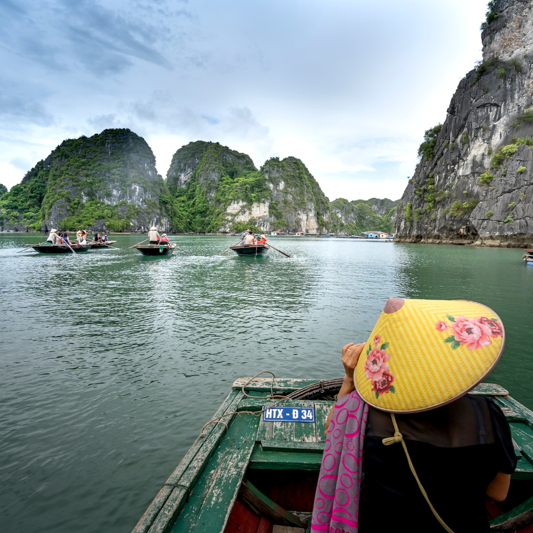 A woman wearing a traditional Vietnamese conical hat sits on a wooden boat in Ha Long Bay, with limestone cliffs and other rowboats in the background. A woman wearing a traditional Vietnamese conical hat sits on a wooden boat in Ha Long Bay, with limestone cliffs and other rowboats in the background.