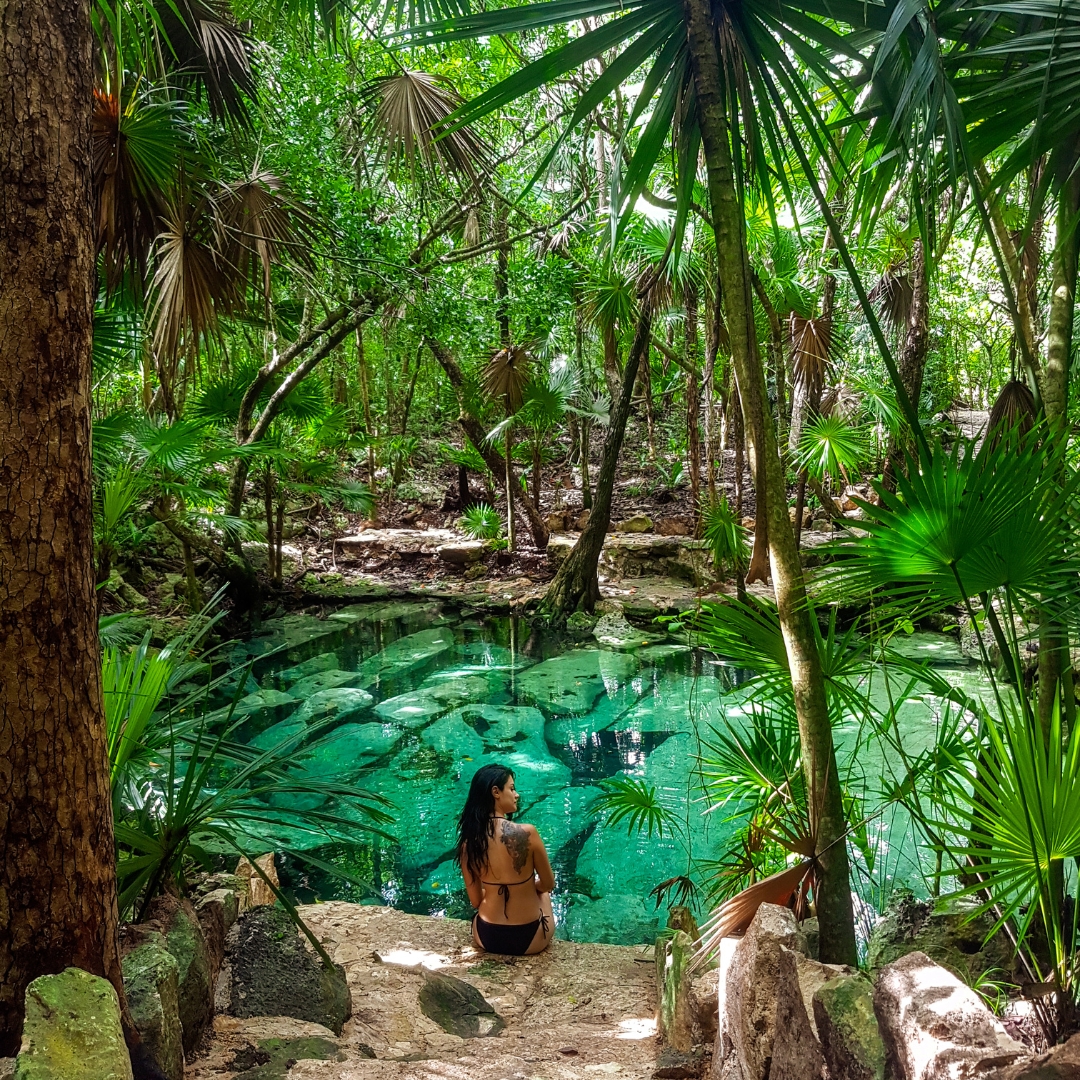 A woman sits at the edge of a crystal-clear cenote surrounded by lush green jungle in Yucatán, Mexico. A woman sits at the edge of a crystal-clear cenote surrounded by lush green jungle in Yucatán, Mexico.