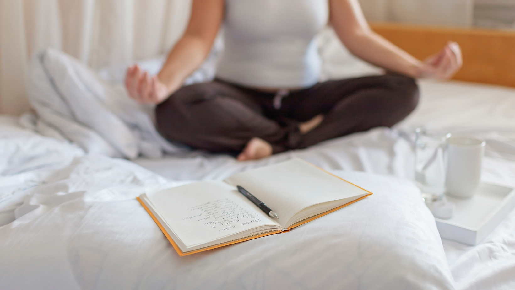 A person is sitting cross-legged on a bed, meditating with hands in a mudra position. In front of them on the bed is an open journal with handwritten notes and a pen placed on top. Nearby, there is a tray with a glass of water and a white mug. The scene suggests a peaceful morning routine involving meditation and journaling for mindfulness and self-reflection.