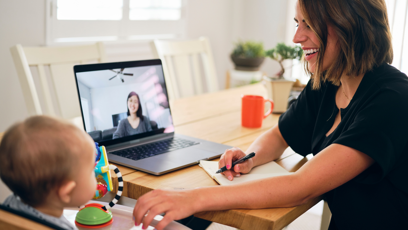 A woman is working from home, sitting at a table with a laptop in front of her. She is smiling and writing in a notebook while having a video call with a colleague visible on the laptop screen. A baby is seated nearby in a high chair, playing with toys. The scene highlights the balance of remote work and parenting, showcasing a modern approach to work-life integration. This image is ideal for topics related to remote work, work-from-home tips, and balancing professional and personal life.