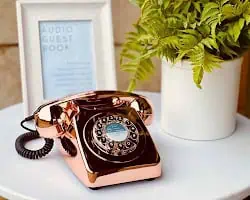 A vintage rotary telephone with a shiny copper finish sits on a white table alongside a potted plant and a framed sign, elegantly doubling as a unique wedding audio guestbook. A vintage rotary telephone with a shiny copper finish sits on a white table alongside a potted plant and a framed sign, elegantly doubling as a unique wedding audio guestbook.