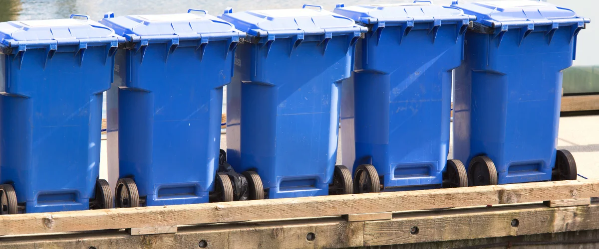 Row of blue trash bins lined up outdoors near the water.