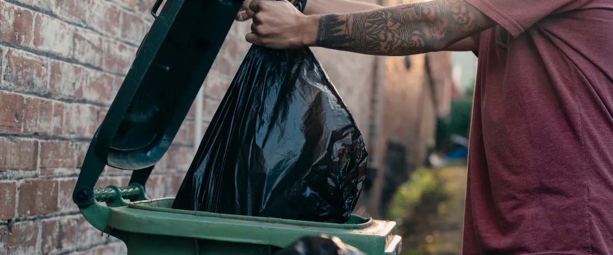 Person lifting the lid of a green garbage bin and placing a black trash bag inside.