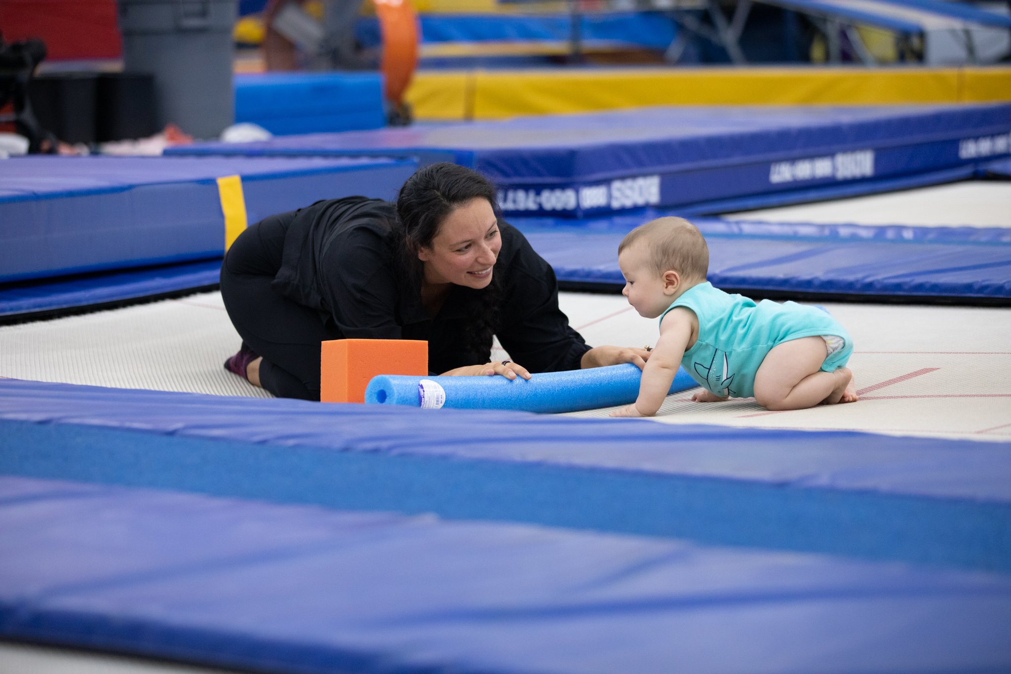 therapist in crouched position holding a noodle in front of a infant crawling on the trampoline