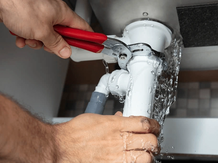 a person's hand using a wrench to fix a leaking white plumbing pipe under a sink, water spraying out.