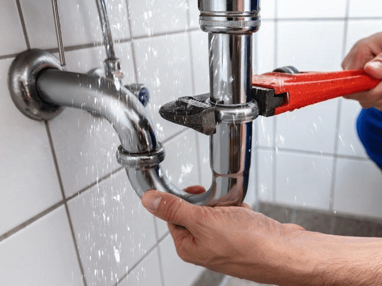 a plumber uses a wrench to fix a leaking pipe under a sink, with water spraying from the joint.