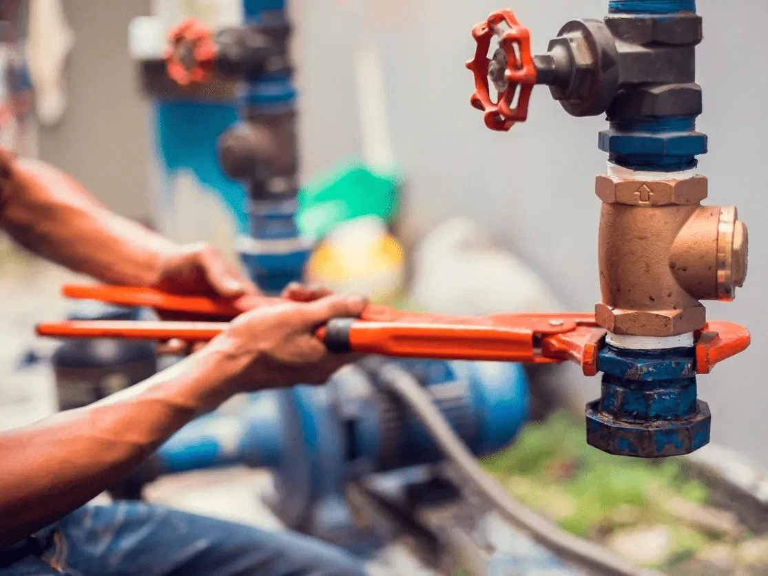 a close-up of a person using large red wrenches to adjust a blue and bronze plumbing valve in an industrial setting.
