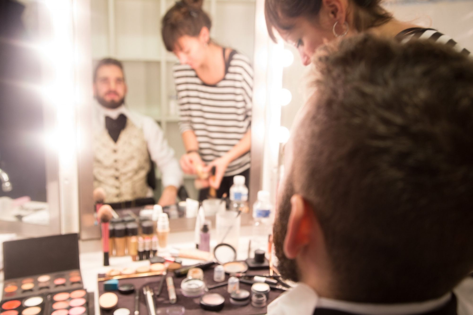 Mirror reflection of a man in light waistcoat and a makeup artist. There are stage lights around the mirror and the image is slightly blurred