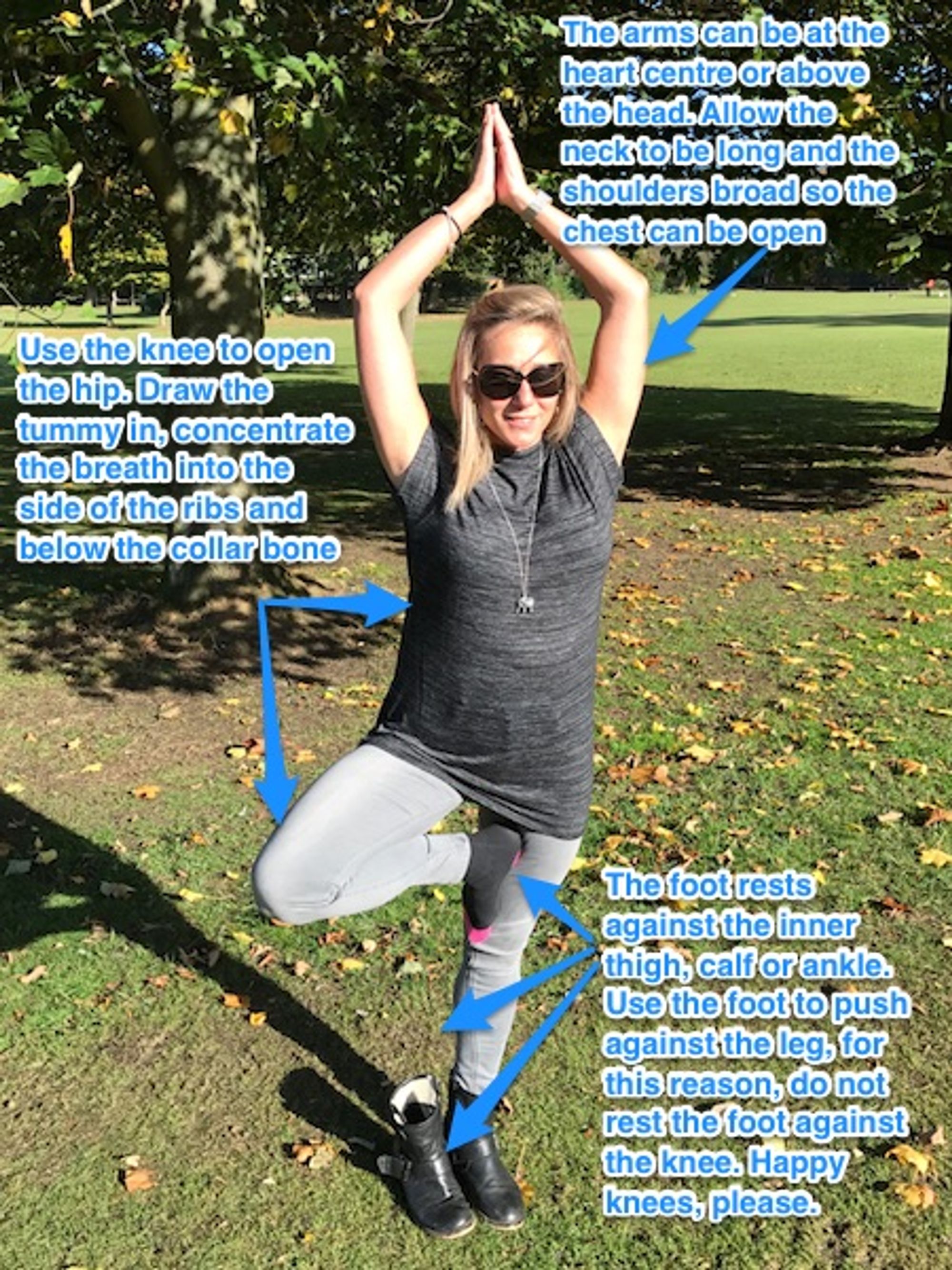 Blonde, long-haired woman in grey jeans and black shirt has taken off one black boot and rested her right foot against her left calf, standing in Tree yoga pose in front of a tree trunk, standing on green grass Blonde, long-haired woman in grey jeans and black shirt has taken off one black boot and rested her right foot against her left calf, standing in Tree yoga pose in front of a tree trunk, standing on green grass