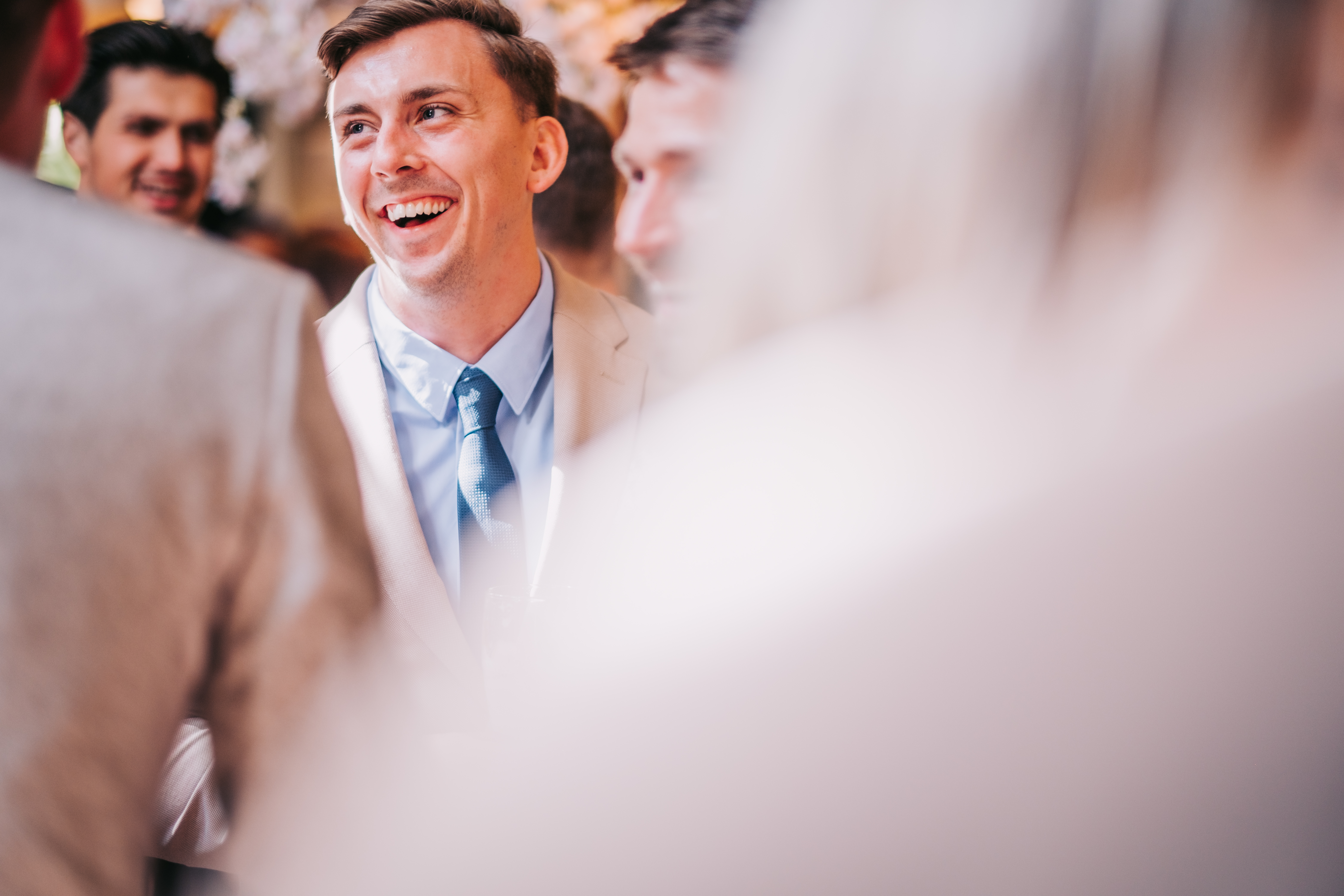 Wedding guest in beige suit laughing during the reception, surrounded by soft foreground blur. Wedding guest in beige suit laughing during the reception, surrounded by soft foreground blur.