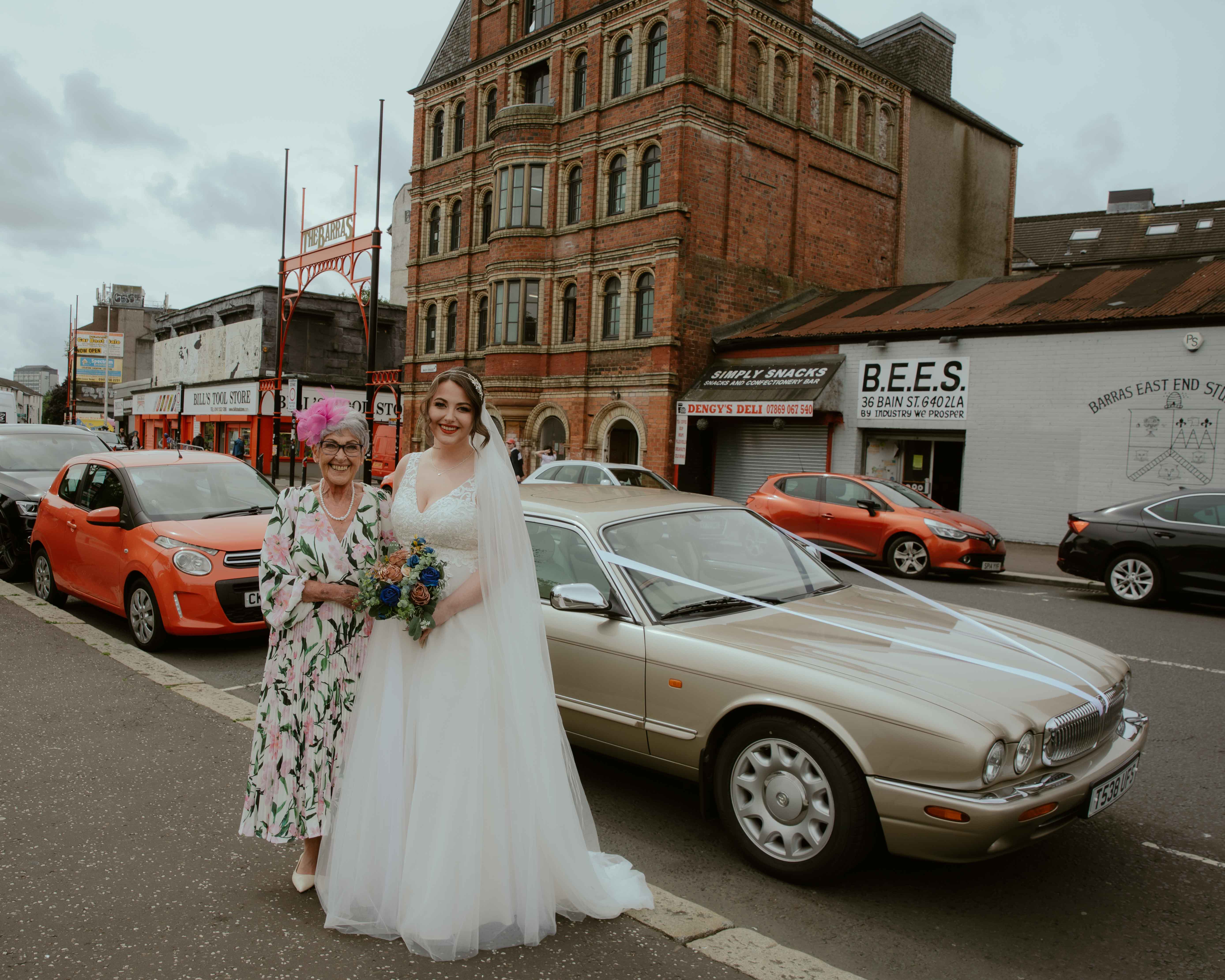 Bride holding her bouquet in the centre with mother of the bride to her left and wedding car behind her. East End, Glasgow wedding. Bride holding her bouquet in the centre with mother of the bride to her left and wedding car behind her. East End, Glasgow wedding.