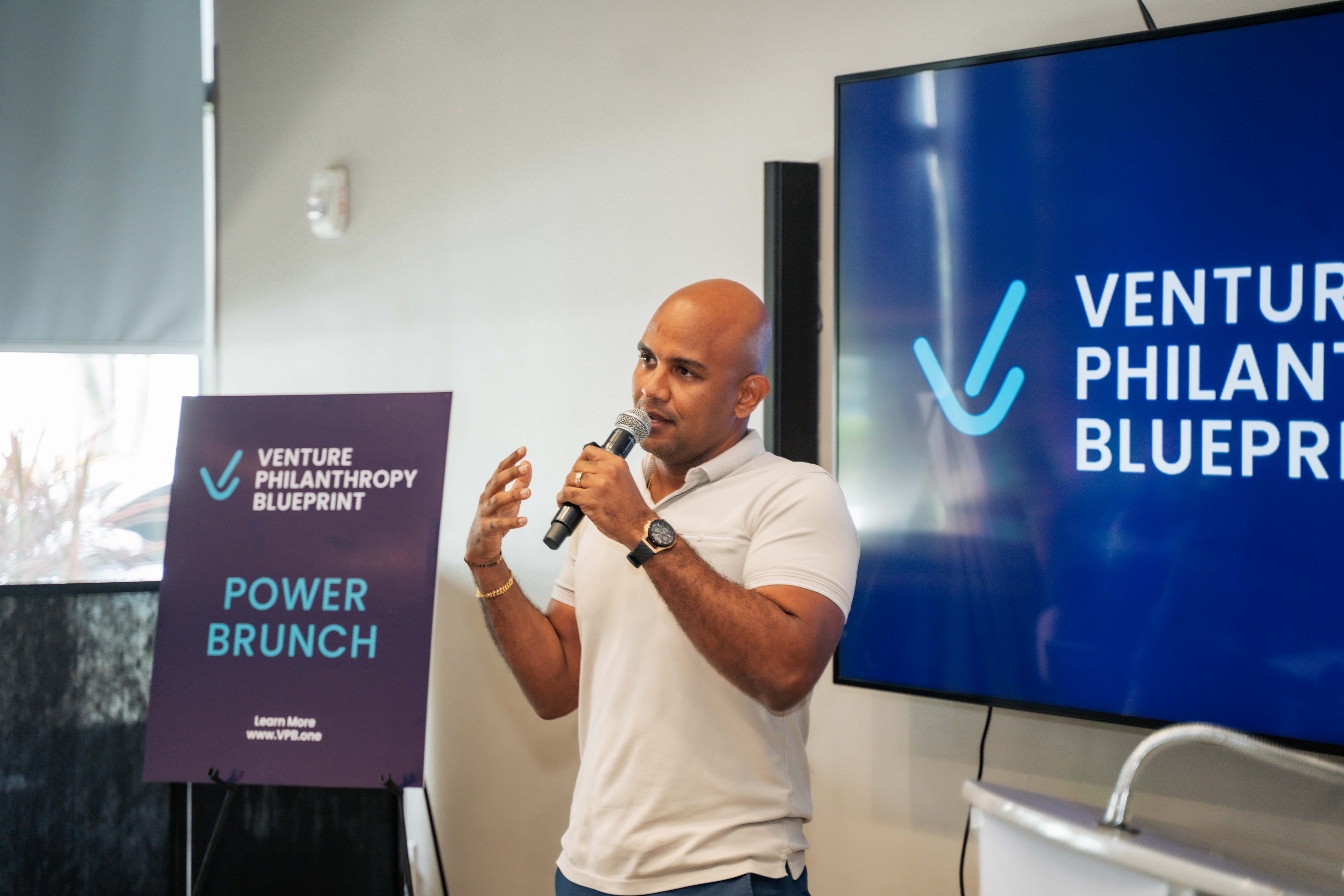 Kirk-Anthony Hamilton speaks into a microphone at the Venture Philanthropy Blueprint Power Brunch event in Kingston, Jamaica. Standing beside a branded sign and in front of a large screen displaying the VPB logo, he gestures expressively while engaging the audience in a discussion focused on innovation and impact.