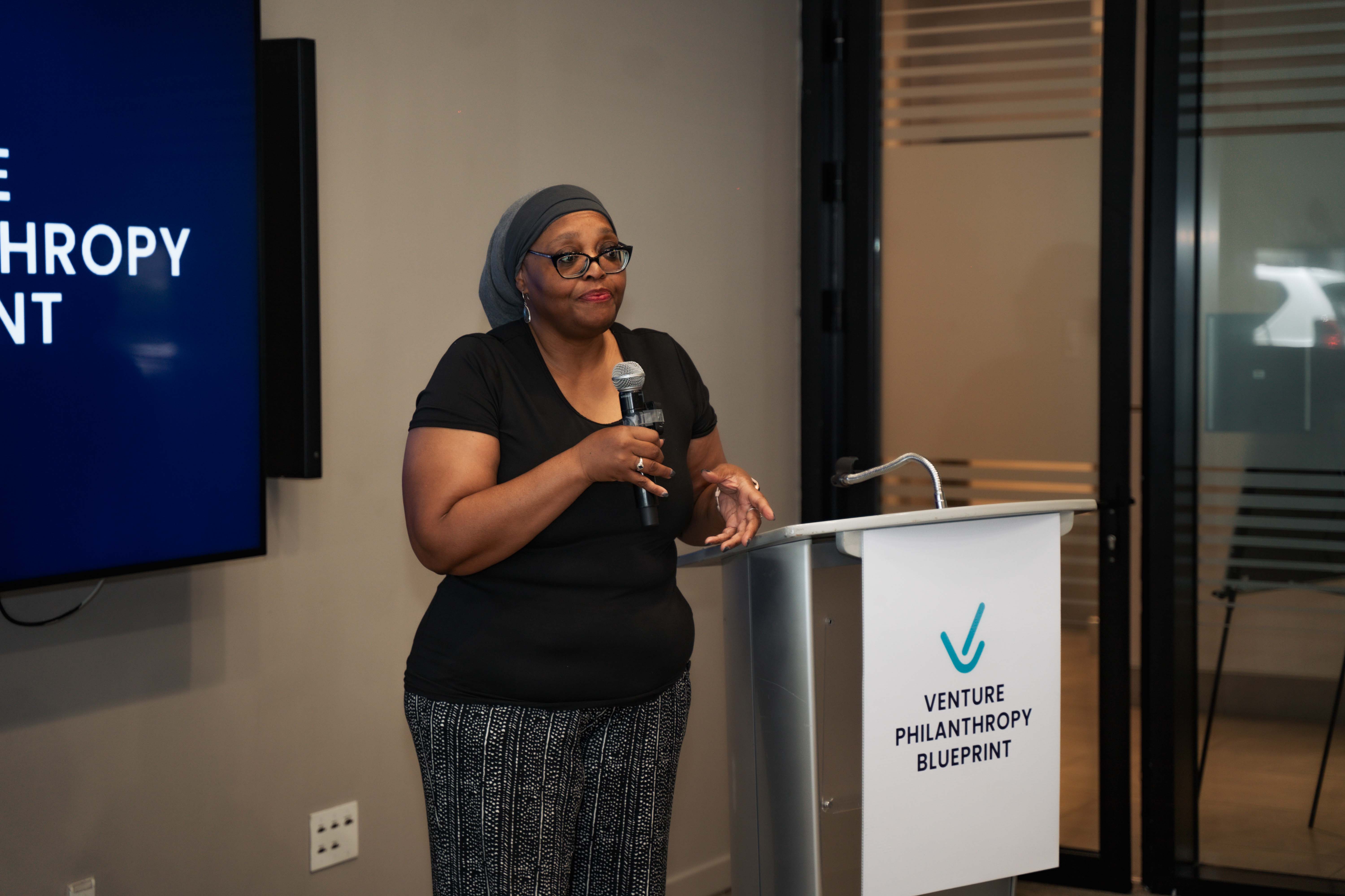 Nedra Daugherty speaks at a podium during the Venture Philanthropy Blueprint Power Brunch event in Kingston, Jamaica. She holds a microphone while addressing the audience, standing beside a branded lectern and in front of a modern, professional setting.